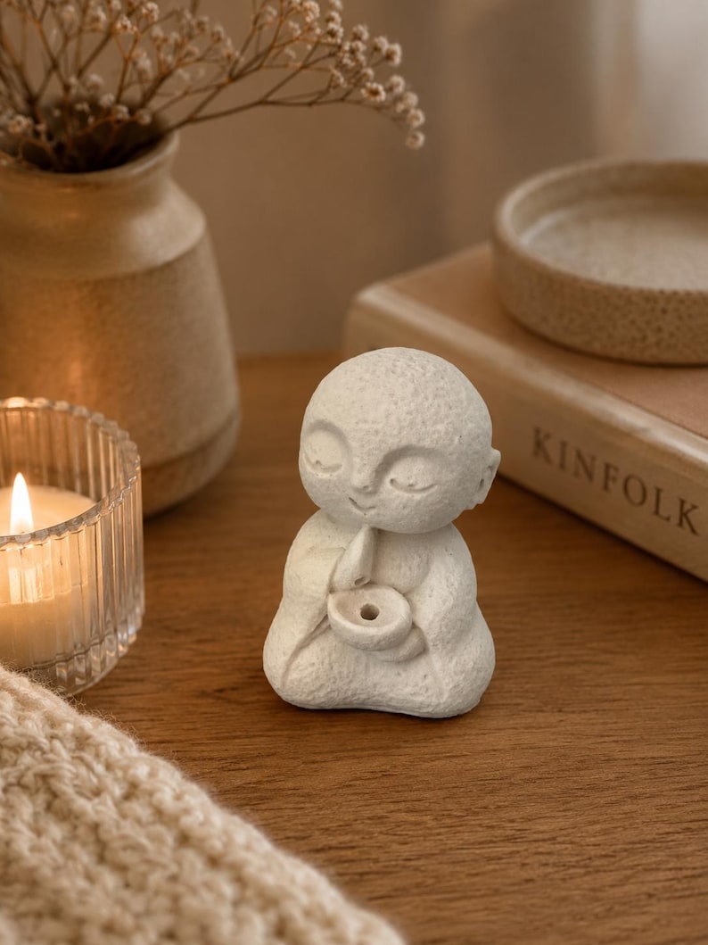 May include: A small, off-white, textured figurine of a seated figure with a smooth, rounded head and closed eyes. The figure is depicted in a meditative pose, holding a small, circular bowl-shaped object with a central hole in its hands.
