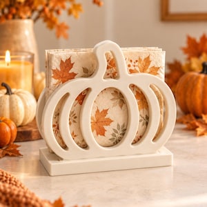 May include: A white pumpkin-shaped napkin holder with a rectangular base, holding a stack of paper napkins printed with a pattern of orange, yellow, and brown autumn maple leaves. The holder features a cutout design resembling a pumpkin silhouette.