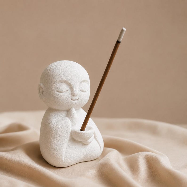 May include: A white matte ceramic figurine of a small person with closed eyes and hands pressed together in a prayer position. The figurine features a small bowl held in the hands, which contains a single brown incense stick pointing upward.
