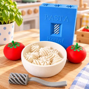 May include: A blue rectangular box labelled PASTA Rotini with a cutout showing white spiral shapes, a white bowl filled with white spiral pasta pieces, two red tomatoes with green stems, and a grey spiral-shaped kitchen tool on a wooden surface.