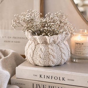 May include: A cream cable-knit basket filled with white flowers. The basket is on top of a stack of books. A lit candle and a framed print are in the background, creating a cosy, neutral-toned aesthetic. The books read 'The Kinfolk Home' and 'Live Beautiful'.