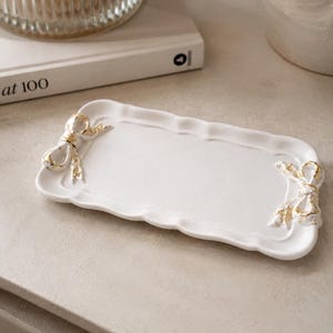 May include: A rectangular white ceramic tray with scalloped edges and a glossy finish. The tray features two decorative three-dimensional bows, one on each short end, accented with distressed gold-coloured paint detailing on the surface.