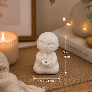May include: A small white matte figurine of a seated person with closed eyes, holding a circular bowl. The figure measures 1.97 inches in height and 1.38 inches in width. The surface has a textured finish, and the figure is depicted in a meditative pose.