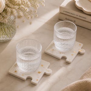 Könnte beinhalten: Zwei klare, geriffelte Glasbecher mit Wasser stehen auf weißen, puzzleförmigen Untersetzern mit Goldflocken. Eine brennende Kerze, eine Vase mit Blumen und Bücher stehen ebenfalls auf dem Tisch. Eine goldene Halskette liegt auf einem dekorativen Teller.