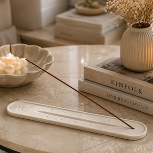 May include: A white, elongated, oval-shaped ceramic incense holder featuring a star emblem at one end and a central groove with small circular indentations along its length, holding a single thin brown incense stick angled upwards.