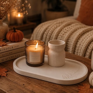 Puede incluir: Una bandeja ovalada blanca con una impresión de hoja de helecho, una vela de cristal ámbar estriado con etiqueta Pumpkin Spice y un pequeño jarrón de cerámica blanca estriada. La vela está encendida sobre una superficie de madera.