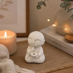 White monk incense holder styled on a wooden table beside a candle, books, and dried flowers in a cozy Scandinavian-inspired home decor scene.