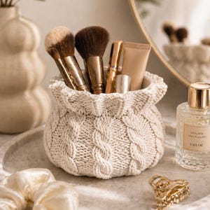 Peut inclure: Un panier en tricot crème rempli de pinceaux de maquillage, de tubes et de contenants. Un flacon de parfum, une pince à cheveux dorée et un chouchou en satin sont posés sur un plateau en marbre. La scène se déroule dans un espace bien éclairé aux tons neutres.