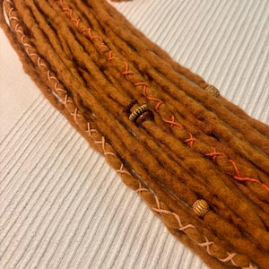 Könnte beinhalten: Ein Bündel handgefertigter, orangefarbener Dreadlocks. Die Dreads sind mit orangefarbenen und beigefarbenen Fadenakzenten und Holzperlen verziert. Im Hintergrund befinden sich ein paar orangefarbene Blumen. Die Dreads liegen auf einer weißen, strukturierten Oberfläche.