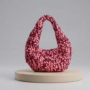 Op de afbeelding: Een kleine, halvemaanvormige handtas gemaakt van dik, gemêleerd garen in tinten diep bordeaux en zacht pastelroze. De tas heeft een doorlopend, geïntegreerd handvatontwerp met een dicht, getextureerd haakpatroon over het gehele oppervlak.