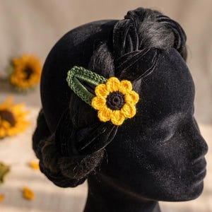 May include: A yellow and black crochet sunflower hair clip with a green leaf backing is displayed on a black mannequin head. The sunflower clip is a bright, cheerful accessory. The background includes other crochet sunflowers.