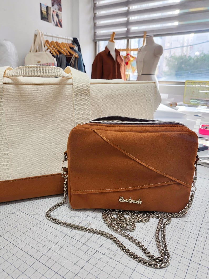 Może przedstawiać: Brązowa torba na ramię ze srebrnym łańcuszkiem i napisem "Handmade" w srebrnym kolorze. Torba ma geometryczny wz&oacute;r i zapięcie na zamek błyskawiczny. Widoczna jest r&oacute;wnież kremowa torba typu tote z brązowymi akcentami.