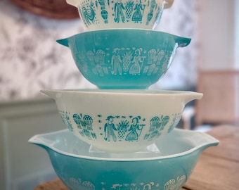 Puede incluir: Una pila de cuatro cuencos apilables con asas, que alternan entre blanco y azul turquesa. Cada cuenco presenta un estampado gráfico repetitivo en blanco o turquesa de figuras, piñas y tallos de trigo alrededor de la superficie exterior.
