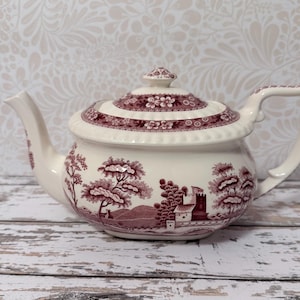 May include: A cream-colored ceramic teapot featuring a maroon transferware pattern of a landscape with trees and a building. The lid has a matching floral border and a small knob handle. The teapot includes a curved spout and a decorative handle.