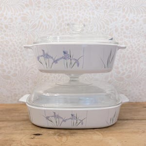 May include: Two white rectangular ceramic baking dishes with clear glass lids stacked vertically. Each dish features a delicate floral pattern of purple irises with green stems and leaves painted along the sides and integrated handles.