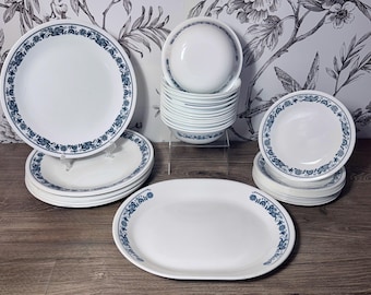 May include: A collection of white dinnerware featuring a blue floral pattern along the rims. The set includes several round dinner plates, a stack of small bowls, a stack of smaller plates, and one large oval serving platter, all matching in design.