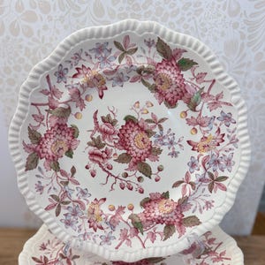 May include: A white ceramic plate with a scalloped edge featuring a detailed floral pattern in shades of deep pink, muted green, and light blue. The design includes clusters of chrysanthemum-like flowers, small buds, and delicate leafy stems.