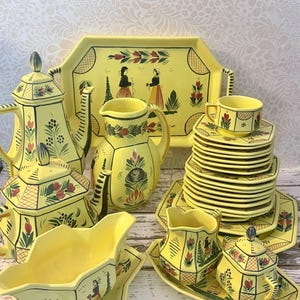 May include: A yellow ceramic tea and dinnerware set featuring hand-painted floral motifs and figures in traditional attire. The collection includes a teapot, a pitcher, a gravy boat, a sugar bowl, a creamer, a large serving tray, and stacked plates.