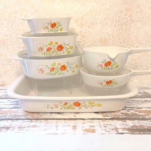 May include: A collection of white ceramic cookware featuring a floral pattern of orange poppies, yellow blossoms, and green foliage. The set includes three rectangular baking dishes with glass lids, a saucepan, a skillet, and a large rectangular tray.