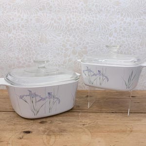 May include: Two white rectangular glass casserole dishes with clear glass lids. Each dish features a delicate botanical illustration of purple iris flowers with green stems and leaves painted on the exterior sides of the white glass body.