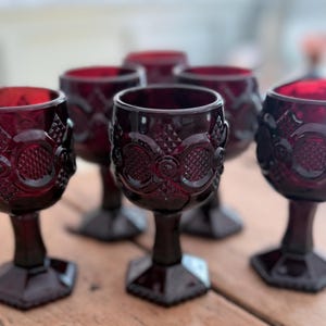 May include: Set of six ruby red goblets. Each goblet features an ornate, embossed design around the bowl and a faceted base. The stem is a dark red color. The goblets are arranged on a wooden surface.