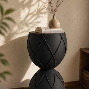 Könnte beinhalten: Ein schwarzer Beistelltisch mit einem einzigartigen geometrischen Design. Der Tisch hat eine runde Ober- und Unterseite, die durch einen schmaleren Mittelteil verbunden sind. Eine kleine Vase mit getrockneten Stielen und zwei Büchern stehen oben. Der Tisch ist ca. 60 cm hoch.