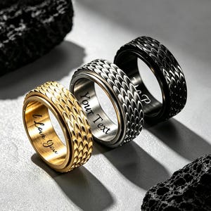 May include: Three metal rings featuring a textured scale pattern on the exterior. The gold-toned ring is engraved with the text I Love You, the silver-toned ring with Your Text, and the black ring with the partial text P on the inner bands.