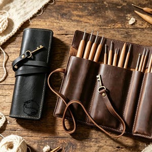 May include: A dark brown leather roll-up case containing multiple wooden crochet hooks, secured with a leather strap and a brass-colored key charm. Beside it sits a matching black leather case with a similar key charm and a small embossed logo.