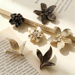 May include: A selection of decorative floral and leaf-shaped hardware in metallic finishes. The collection includes bronze, gold, and black tones, with detailed petal and leaf textures. These pieces are likely cabinet knobs or drawer pulls.