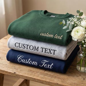 Peut inclure: Une pile de trois sweat-shirts à col rond en vert foncé, gris chiné et bleu marine. Chaque sweat-shirt présente un texte personnalisé brodé sur la poitrine. Le sweat-shirt vert du haut a un texte cursif, le gris un texte en bloc et le bleu marine une écriture.