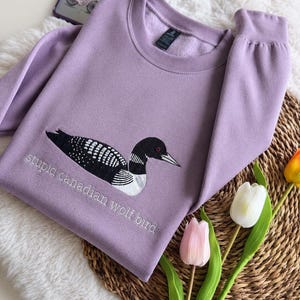 Pode incluir: Uma camisola de gola redonda cor de lavanda com um bordado de um mergulh&atilde;o em preto e branco. Por baixo da ave, o texto "stupid canadian wolf bird" est&aacute; bordado com fio branco. A camisola tem mangas compridas e uma gola canelada.