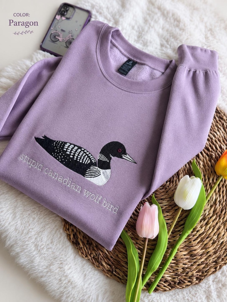 Pode incluir: Uma camisola de gola redonda cor de lavanda com um bordado de um mergulh&atilde;o em preto e branco. Por baixo da ave, o texto "stupid canadian wolf bird" est&aacute; bordado com fio branco. A camisola tem mangas compridas e uma gola canelada.