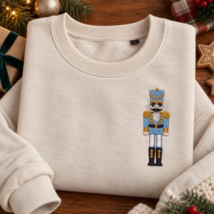May include: A beige crewneck sweatshirt featuring a small, detailed embroidery of a nutcracker on the left chest. The nutcracker is depicted in light blue, gold, and black thread, wearing a tall hat, a uniform with gold accents, and black boots.