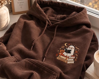 May include: A dark brown hooded sweatshirt featuring an embroidered graphic on the chest. The design depicts a white ghost wearing round glasses, reading a book while sitting on a stack of three books, surrounded by a pumpkin, stars, and leaves.