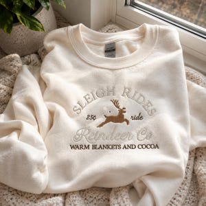 May include: A cream-colored crewneck sweatshirt featuring brown embroidery on the chest. The design includes a leaping reindeer, the text "SLEIGH RIDES", "25&cent; ride", "Reindeer Co.", and "WARM BLANKETS AND COCOA" in a mix of block and script fonts.