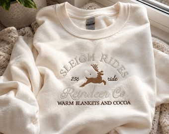 May include: A cream-coloured crewneck sweatshirt featuring brown embroidery on the chest. The design includes a leaping reindeer, the text "SLEIGH RIDES", "25¢ ride", "Reindeer Co.", and "WARM BLANKETS AND COCOA" in a mix of block and script fonts.