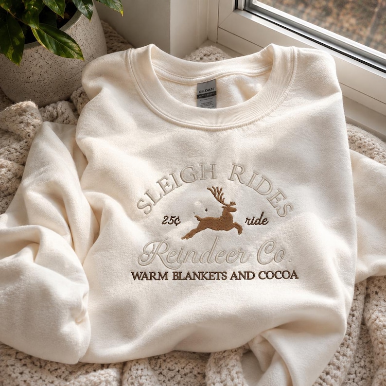 May include: A cream-colored crewneck sweatshirt featuring brown embroidery on the chest. The design includes a leaping reindeer, the text "SLEIGH RIDES", "25&cent; ride", "Reindeer Co.", and "WARM BLANKETS AND COCOA" in a mix of block and script fonts.
