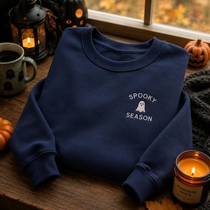 Könnte beinhalten: Ein marineblaues Sweatshirt mit Rundhalsausschnitt und weißem Sticktext, der SPOOKY SEASON über und unter einer kleinen weißen Geistergrafik auf der linken Brustseite zeigt.