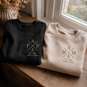 Könnte beinhalten: Ein schwarzes Sweatshirt und ein beiges Sweatshirt, jeweils mit einem kreuzförmigen Design mit dem Text PAPA oder MAMA, gefolgt von Est. 2021 und den Namen Luca und Elias, gedruckt in einer Serifenschrift auf der linken Brustseite.