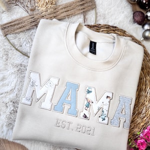 Könnte beinhalten: Ein beigefarbenes Sweatshirt mit Rundhalsausschnitt und dem Wort MAMA in großen Buchstaben im College-Stil, die mit verschiedenen Mustern und Farben wie Hellblau und Charakter-Prints versehen sind. Unter MAMA ist der Text EST. 2021 in Beige gestickt.