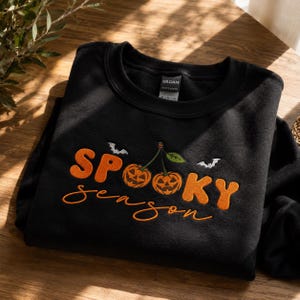 Puede incluir: Una sudadera negra de cuello redondo con texto bordado en naranja que dice SPOOKY SEASON. El diseño incluye dos calabazas de Halloween naranjas con tallos y hojas verdes, dispuestas como cerezas, acompañadas de dos pequeños murciélagos blancos.