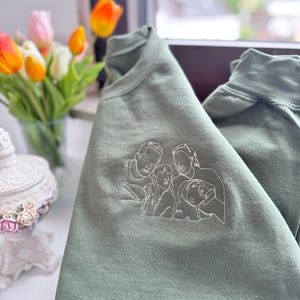May include: An olive green crewneck sweatshirt featuring a white line art embroidery of a family of four on the chest. The design depicts the outlines of two adults and two children positioned closely together in a portrait style illustration.