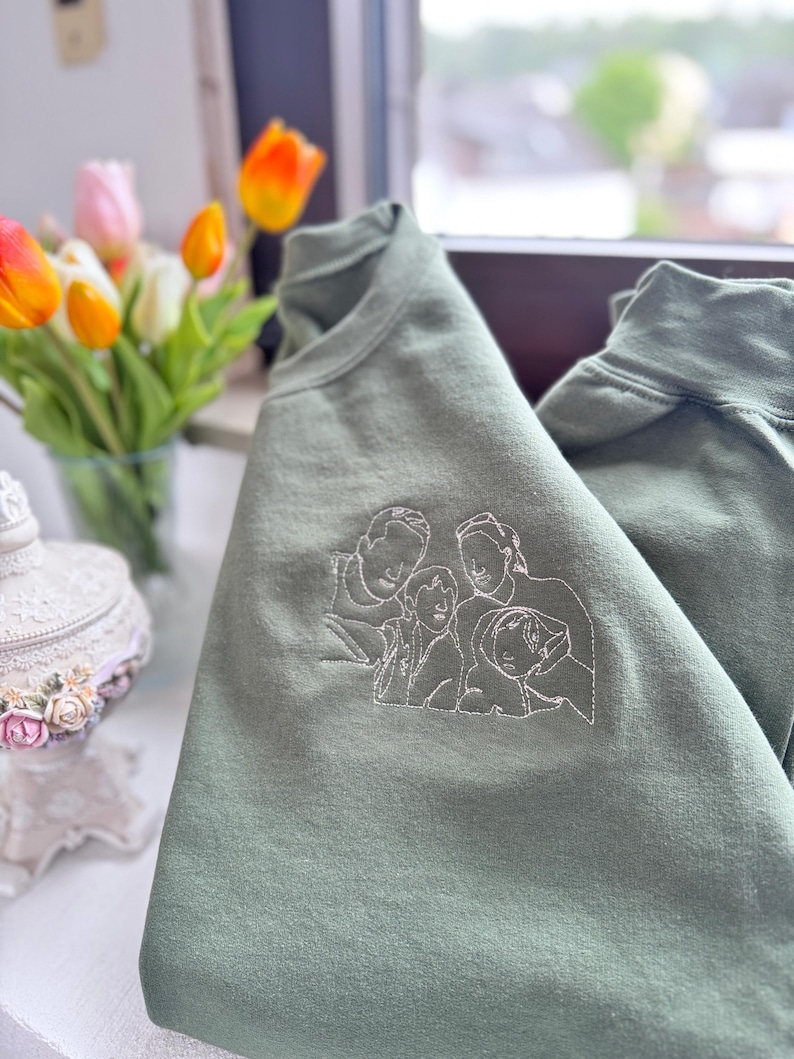 May include: An olive green crewneck sweatshirt featuring a white line art embroidery of a family of four on the chest. The design depicts the outlines of two adults and two children positioned closely together in a portrait style illustration.