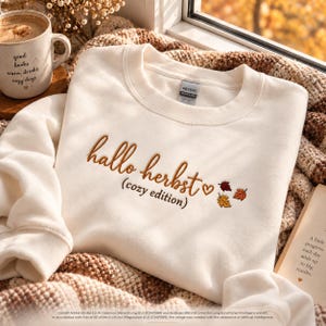 May include: A cream-coloured crewneck sweatshirt featuring the embroidered brown text "hallo herbst (cozy edition)" with a small heart and three colourful autumn leaf icons in shades of orange, red, and yellow on the front chest.