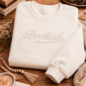 Könnte beinhalten: Ein cremefarbenes Sweatshirt mit Rundhalsausschnitt, auf dessen Brustmitte das Wort Bookish in passender Garnfarbe gestickt ist. Das Kleidungsstück hat lange Ärmel mit gerippten Bündchen sowie einen gerippten Halsausschnitt aus einfarbigem Stoff.