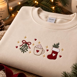 Könnte beinhalten: Ein cremefarbenes Sweatshirt mit Rundhalsausschnitt und gestickten Weihnachtsmotiven, darunter ein Mistelzweig mit roter Schleife, eine Tasse heiße Schokolade mit Sahne und ein roter Weihnachtsstrumpf, verziert mit kleinen roten Herzen und goldenen Sternen.