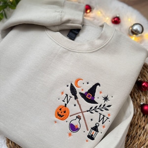 Könnte beinhalten: Ein beigefarbenes Sweatshirt mit Rundhalsausschnitt und einer Stickerei auf der Brust. Das Motiv zeigt einen Kürbis, einen Hexenhut, einen Besen, eine Flasche, eine Laterne, eine Mondsichel, Sterne sowie die Buchstaben N und W in Schwarz.