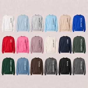 May include: A collection of eighteen crewneck sweatshirts displayed in a grid, each featuring a different solid color including white, ash grey, sport grey, sky blue, sand, royal blue, red, pink, paragon, off white, navy, military green, forest green, stone blue, cocoa, dark heather, black, and charcoal, with the color name printed vertically in black text on the right side of each garment.