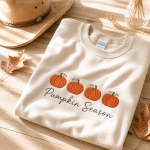 Puede incluir: Una sudadera de cuello redondo color crema con un diseño bordado de cuatro calabazas naranjas con tallos y zarcillos verdes, situadas sobre el texto negro en cursiva "Pumpkin Season" centrado en la zona del pecho.