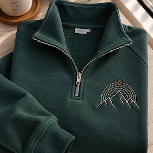 May include: A dark green quarter-zip sweatshirt featuring a silver metal zipper and a ribbed collar. An embroidered design on the chest depicts a mountain range with concentric semicircular lines above it and a small crescent moon at the top.